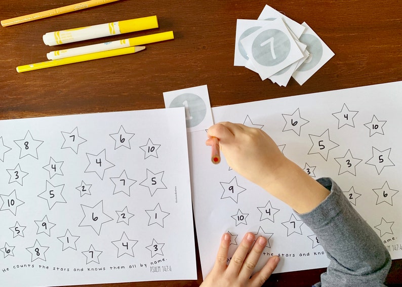 May include: A child's hand places a number sticker on a star-shaped worksheet. The worksheet has stars with numbers inside and the text "He counts the stars and knows them all by name. Psalm 147:4".