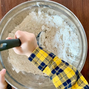 May include: A child's hand uses a black and silver pastry cutter to mix dry ingredients in a silver bowl. The bowl is filled with white flour and other dry ingredients.