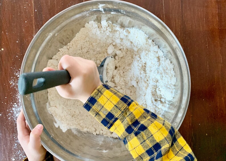 May include: A child's hand uses a black and silver pastry cutter to mix dry ingredients in a silver bowl. The bowl is filled with white flour and other dry ingredients.