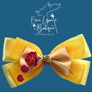 Puede incluir: Un lazo para el cabello amarillo y dorado con una rosa roja y detalles de pétalos rosas. El lazo presenta un centro de purpurina dorada y está sobre un fondo azul. El texto "Once Upon a Bootique Accessories" está en la parte superior.