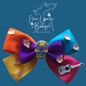May include: A vibrant hair bow with a Day of the Dead design. The bow incorporates orange, yellow, purple, blue, and pink ribbons. A skull-shaped decoration is at the centre, and a small guitar charm is attached to the pink ribbon. The text "Once Upon a Bootique Accessories" is also visible.