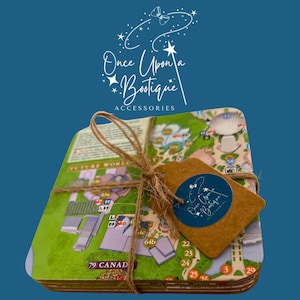 Può includere: Una pila di sottobicchieri quadrati con una mappa colorata di un parco a tema, legati insieme con spago marrone. Un'etichetta di carta marrone è attaccata allo spago, mostrando un adesivo blu circolare con il testo bianco Once Upon a Bootique Accessories.