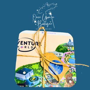 May include: A stack of square coasters featuring a colorful map illustration of a theme park with the text Adventure World and Adventure Way. The coasters are tied together with brown twine and include a small brown paper tag with a blue circular logo.