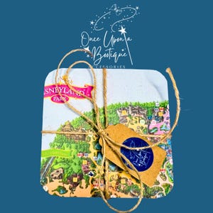 Puede incluir: Un posavasos cuadrado con un mapa colorido del parque Disneyland, atado con cuerda marrón y una pequeña etiqueta de papel marrón. La etiqueta muestra una pegatina circular azul con el texto blanco Once Upon a Boutique Accessories.