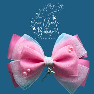 Puede incluir: Un lazo para el pelo grande rosa y azul con un dije de varita de estrella. El lazo tiene un diseño en capas con un centro brillante y está adornado con perlas rosas. El texto "Once Upon a Boutique" está en el fondo.