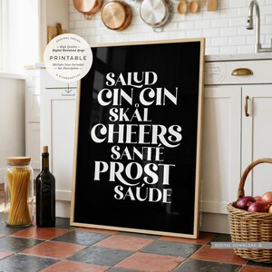 May include: A rectangular framed wall art piece with a black background and white typography. The text reads: SALUD, CIN CIN, SKÅL, CHEERS, SANTÉ, PROST, SAÚDE. A circular white badge in the top left corner contains the text: ORIGINAL DESIGN, High Quality, Digital Download Jpegs, PRINTABLE, Multiple Sizes Included, See Description, STARSANDTYPE.