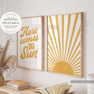 May include: Two framed prints with a retro theme. One features the text "Here comes the Sun" in yellow on a white background. The other print displays a yellow sunburst design. Both are in wooden frames.