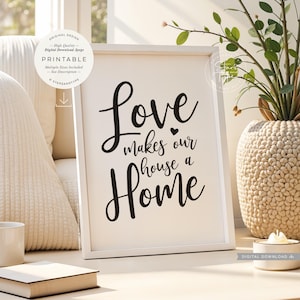 Może przedstawiać: Biała oprawiona grafika z napisem "Love makes our house a Home" czcionką. Grafika jest wyświetlana na jasnej powierzchni, z dekoracyjnym wazonem i małą świecą w pobliżu. Widoczne są również słowa "Original Design" i "Printable".