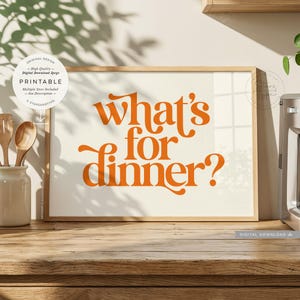 May include: A rectangular wooden frame containing a white print with the orange text "what's for dinner?" in a stylized font. The frame is positioned on a wooden surface next to a white ceramic container holding two wooden spoons.