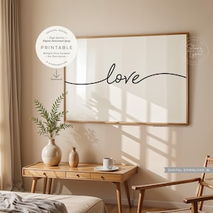 May include: A printable wall art print featuring the word "love" written in a cursive font. The print is framed in a gold frame and is hanging on a beige wall. The print is in a living room setting with a wooden table and chair.