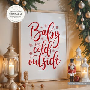 Op de afbeelding: Een ingelijste witte print met de woorden "Baby it's cold outside" in rode letters. De print is versierd met sneeuwvlokken. De afbeelding maakt deel uit van een kerstthema, met ornamenten en een kaarslantaarn.
