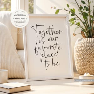 Op de afbeelding: Een witte ingelijste print met de tekst "Together is our favorite place to be" in zwart script. Het frame staat op een licht oppervlak, met een decoratieve vaas en een kleine kaars. De print is een digitale download.