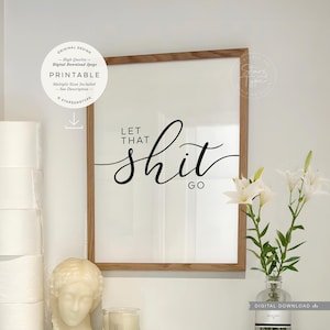 May include: A framed print with the words "LET THAT shit GO" in black script. The print is in a light wood frame and is displayed on a white wall. The image also includes a stack of toilet paper, a bust, and a vase of white flowers.
