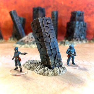 Op de afbeelding: Een 3D-geprint miniatuur van een futuristische, gebroken, grijze en blauwe kolom, met een textuur. Twee miniatuurfiguren, een in blauw en een in grijs, staan ​​naast de kolom.