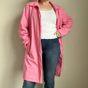 May include: A bright pink trench coat with a collared neckline and long sleeves. The coat is open, revealing a white tank top and blue jeans. The coat has pockets and a classic, longline silhouette.