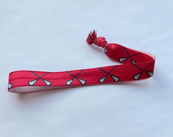 May include: A red elastic hair tie featuring a pattern of crossed lacrosse sticks in black and white. The hair accessory is knotted at one end and lies flat, showcasing the repeating design. The background is a plain white surface.