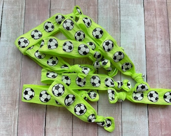 May include: Several neon green hair ties with a repeating pattern of black and white soccer balls. The hair ties are knotted and arranged on a light wood-grain surface.
