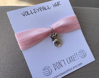 May include: A light pink elastic hair tie featuring a silver-toned metal charm shaped like a volleyball with a heart on top. The hair tie is attached to a white display card with the text VOLLEYBALL HAIR DON'T CARE and volleyball graphics.