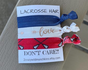 May include: A set of three lacrosse-themed hair ties. One is navy blue, one is white with a "love" graphic, and the third is red with lacrosse stick graphics. The card reads "LACROSSE HAIR DON'T CARE!!"