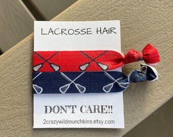 May include: Two red and blue hair ties with a lacrosse stick pattern. The hair ties are on a white card with the text "Lacrosse Hair Don't Care!! 2crazywildmunchkins.etsy.com"