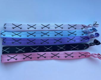 May include: Set of six colorful elastic hair ties with a black criss-cross design. The hair ties are in shades of pink, purple, blue, and white.