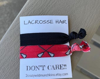 May include: Two hair ties on a white card with the text "LACROSSE HAIR" and "DON'T CARE!!". One hair tie is black, and the other is red with a white lacrosse stick design. The Etsy shop URL is visible.