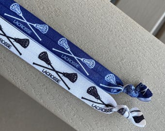 May include: Two elastic hair ties featuring a repeating pattern of crossed lacrosse sticks and the word LACROSSE. One hair tie is dark blue with white graphics, and the other is white with dark blue graphics. Both have a knotted end detail.
