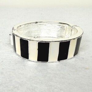 May include: A silver-toned hinged bracelet with a black, white, and cream striped pattern. The bracelet has a rectangular shape with a clasp closure. The design features alternating vertical stripes of black, white, and cream.