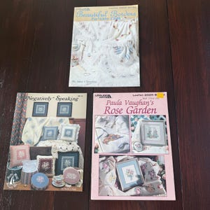May include: Three vintage craft books are displayed on a dark wooden surface. The books feature titles like "Beautiful Borders," "Negatively Speaking," and "Paula Vaughan's Rose Garden." The covers showcase needlework designs.