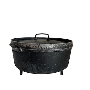 May include: A round black cast iron Dutch oven with a matching lid, featuring a wire handle on top and two side handles. The exterior surface shows signs of wear with silver-toned metallic scuffing along the rim and base, supported by three legs.