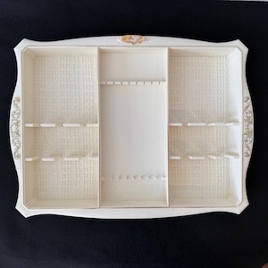 May include: Ivory-colored jewelry organizer with multiple compartments and a decorative gold accent. The organizer has a rectangular shape with curved edges and ornate detailing. It features various sections for storing jewelry.