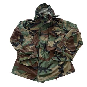 May include: A long-sleeve field jacket featuring a woodland camouflage pattern in shades of green, brown, and black. The jacket includes a hood, a front zipper closure with a buttoned storm flap, two chest pockets with flaps, and adjustable cuffs.