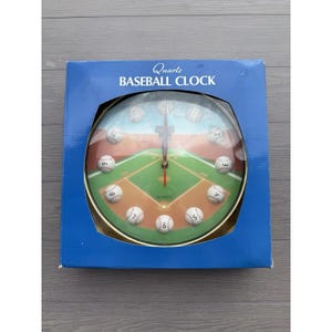 May include: A quartz baseball clock with a baseball field design. The clock face features a green field, brown base paths, and baseballs marking the hours. The clock is in a blue box with the words "Quartz Baseball Clock".