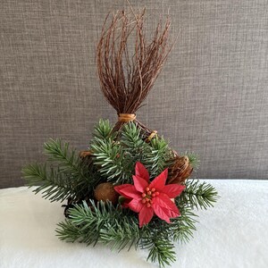 May include: A festive Christmas decoration featuring a cluster of green pine needles, a vibrant red poinsettia, and a bundle of brown twigs. The arrangement includes pine cones and is tied with twine.