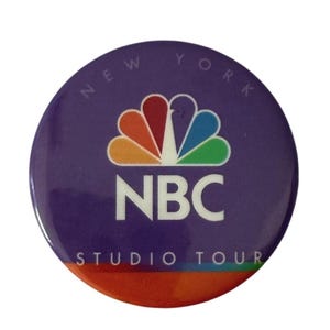 May include: A round purple button with the NBC logo in the center. The logo features a colorful peacock design above the white letters "NBC". The words "NEW YORK" are arched at the top, and "STUDIO TOUR" is at the bottom.
