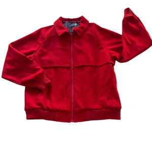 May include: A bright red zip-up jacket featuring a classic collar, long sleeves with ribbed cuffs, and a ribbed hem. The interior lining displays a multi-colored plaid pattern in shades of green, blue, and red. The jacket has a front zipper closure.