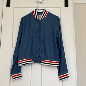 May include: A blue denim bomber jacket with a button-up front. The collar, cuffs, and hem feature white, red, and black stripes. The jacket is hanging on a white hanger against a white door.