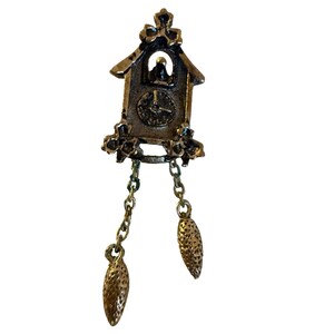May include: A vintage-style gold-toned metal cuckoo clock pendant. The pendant features a clock face, a small bird, and decorative chains with cone-shaped charms. The clock has a bow at the top and floral accents.