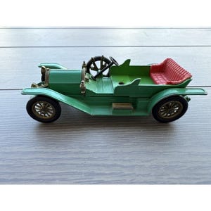 May include: A green vintage toy car with a red seat. The car has black wheels with gold spokes, a black steering wheel, and a detailed front end. The car is a classic model, likely from the early 20th century.