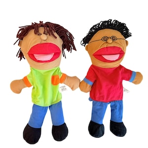May include: Two plush hand puppets with brown skin tones. The left puppet has brown yarn hair, a lime green shirt with an orange pocket, blue pants, and black shoes. The right puppet has black yarn hair, glasses, a red shirt, blue pants, and black shoes.