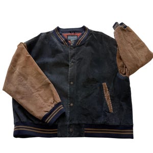 May include: A varsity-style jacket featuring a dark navy blue body and contrasting tan brown sleeves. The garment includes a ribbed collar, cuffs, and hem with navy and tan stripes, a front snap-button closure, and two side pockets with tan trim.