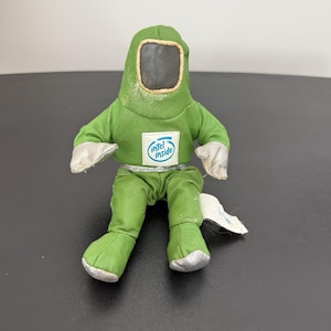 May include: A green plush doll in a hazmat suit with a black face shield and white gloves. The doll has the "Intel Inside" logo on the chest. The doll is sitting on a dark surface.