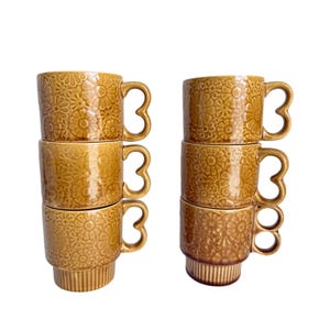 May include: Six stackable ceramic mugs in a warm mustard yellow glaze. Each mug features an embossed floral pattern across the exterior and a distinct heart-shaped handle. The base of each mug has a vertical ribbed texture for secure stacking.