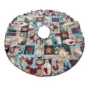May include: A circular tree skirt featuring a woven tapestry pattern of snowmen, red hearts, brown stars, and birds on a light blue background. The text "Snowmen Melt, Memories Last" and "Joy" is printed in dark lettering across the fabric surface.