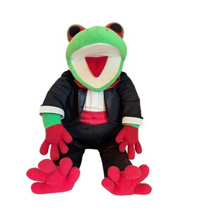 May include: A plush frog toy with green skin, large black eyes, and an open mouth showing a red tongue. The frog is dressed in a black tuxedo jacket with a white shirt front, a red cummerbund, and red gloves on its hands and feet.