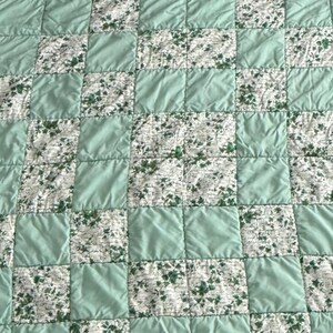 May include: A patchwork quilt featuring a repeating pattern of squares. Some squares are a solid light green, while others display a white background with a green floral design. The quilt has a quilted texture.