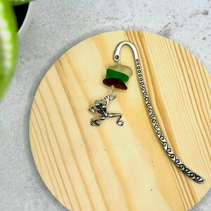 Peut inclure: Signet en argent avec un charme de grenouille et une perle de verre en couches dans des tons de vert, blanc et marron. Le signet est sur une surface en bois.