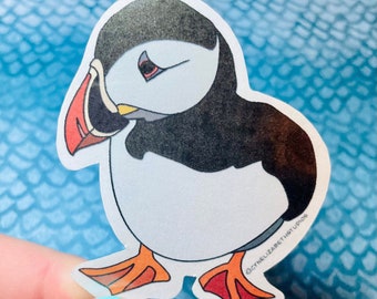 May include: A white and black cartoon sticker of a puffin with an orange beak and feet. The sticker has a white background and the text "@cynelizabethstudios" in the bottom right corner.