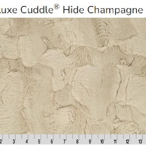 May include: Close-up of a beige faux fur fabric with a textured, wavy pattern. The fabric is labelled "Luxe Cuddle® Hide Champagne".