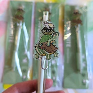 May include: A clear pen with a stack of four turtles on top. The turtles are green, brown, and white with black eyes. The turtles are stacked on top of each other, with the smallest turtle on top. The pen is white with a silver clip.